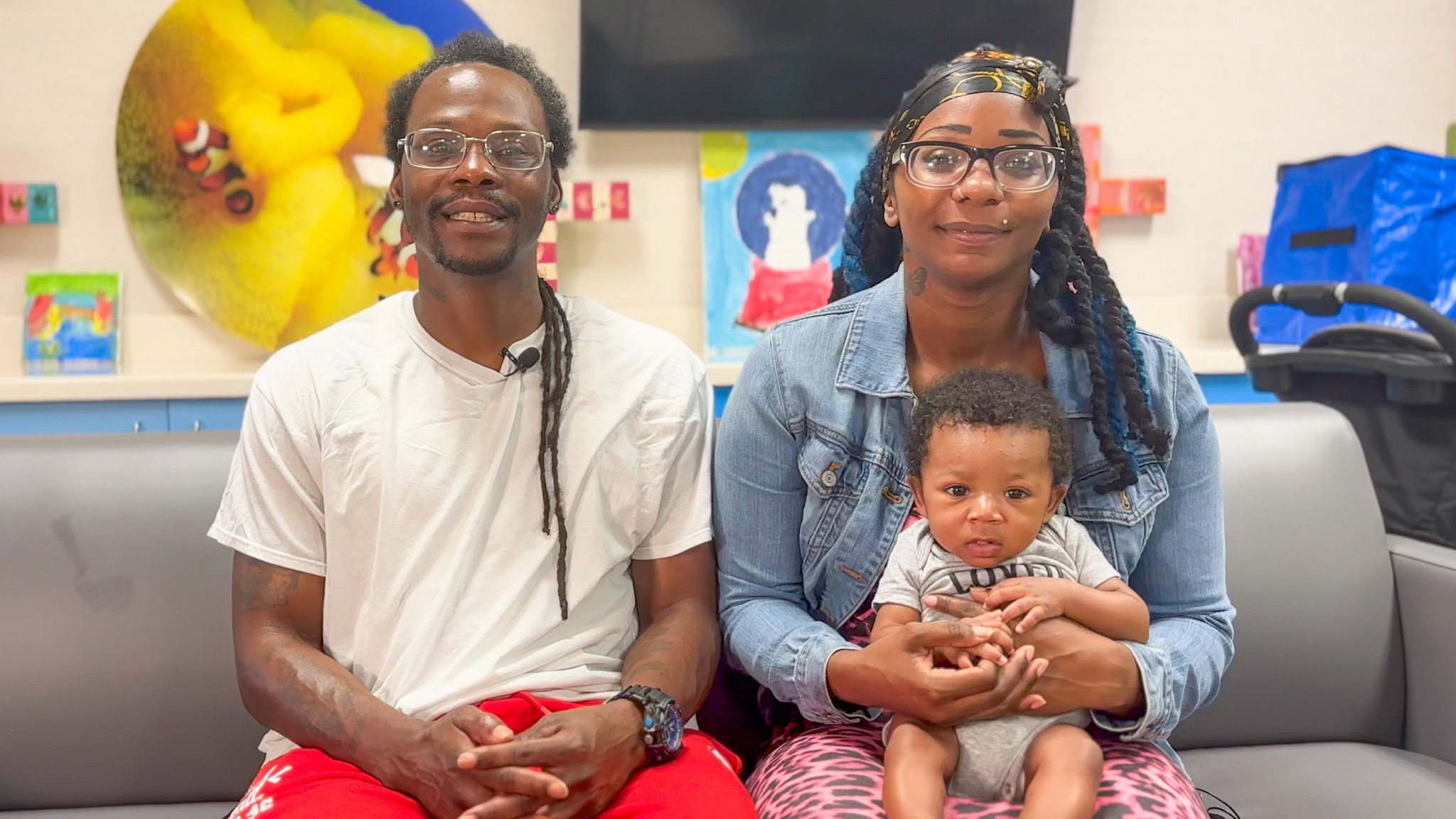 A father, mother, and baby sit on a sofa looking at the camera.